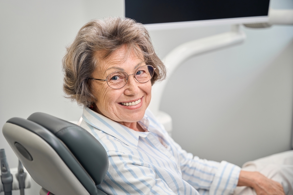 Image of smiling senior woman with snap in dentures, confidently eating an apple in Rochester, NY. The background includes a blurred view of the Rochester skyline. No text on image.